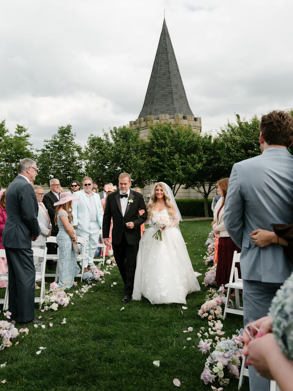 Bride in lace gown walks down flower-lined aisle with groom at castle venue.
