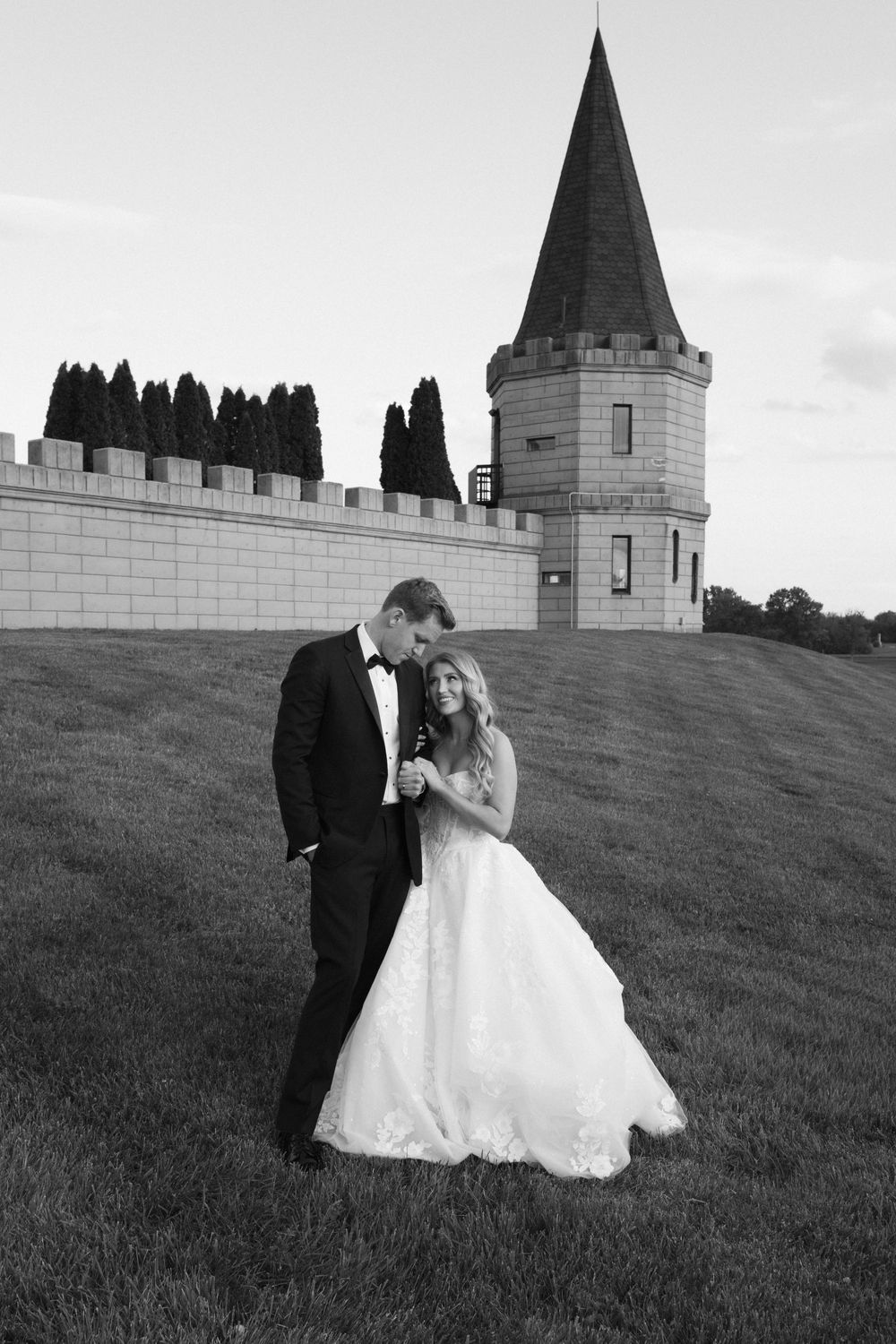 Black and white photo of a bride and groom embracing on a grassy hill beside a castle tower.