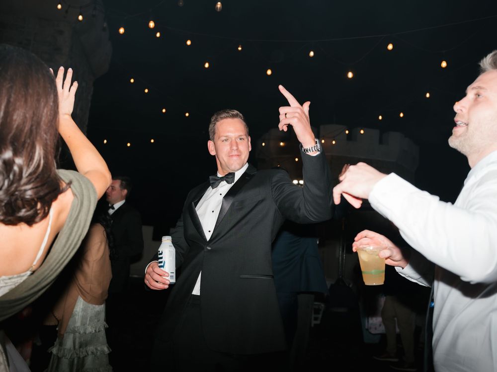 Man in tuxedo dancing at elegant wedding reception under string lights with guests celebrating around him.