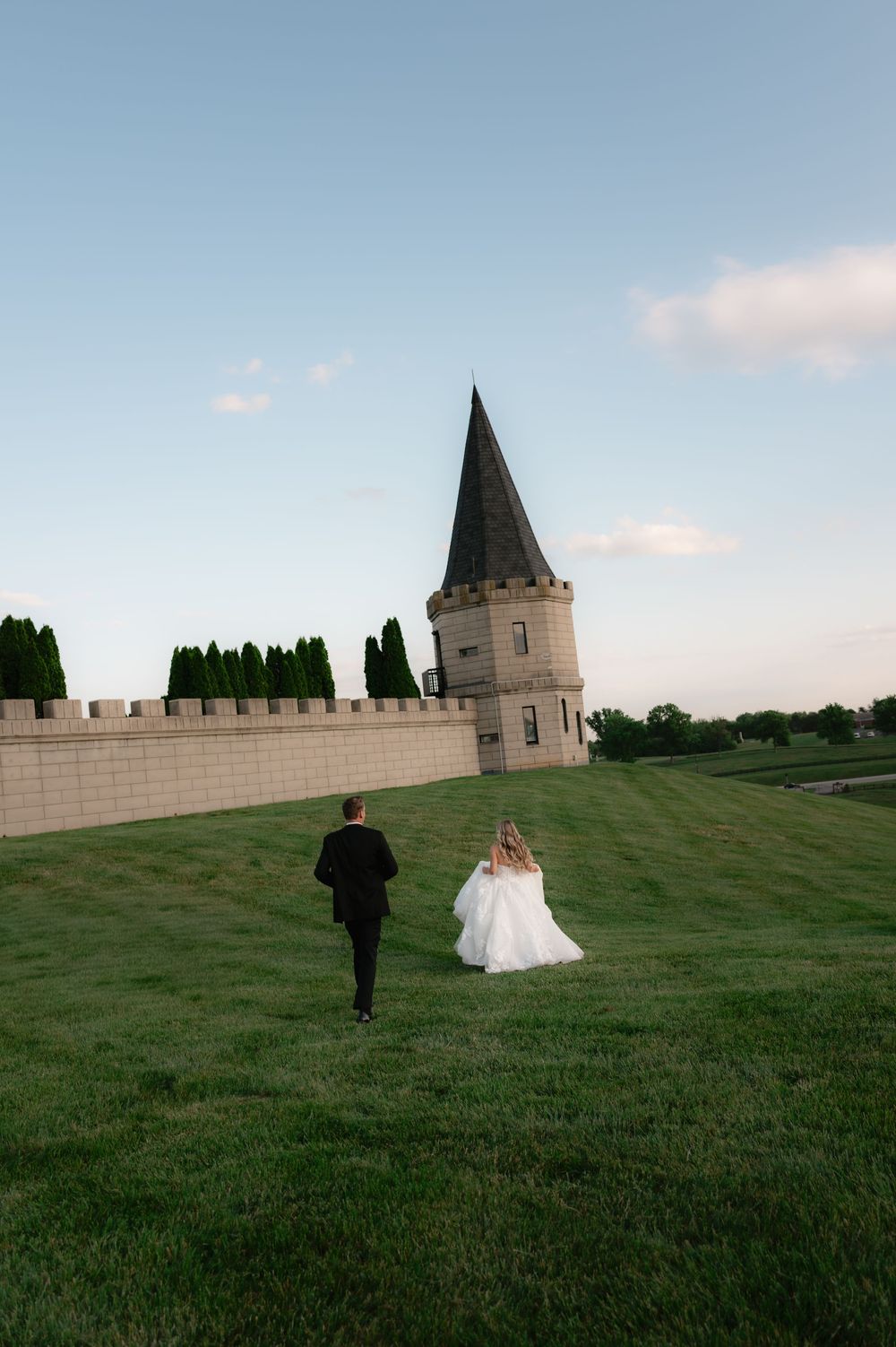A bride and groom walk hand in hand across a lush green lawn toward a stone castle tower at dusk.