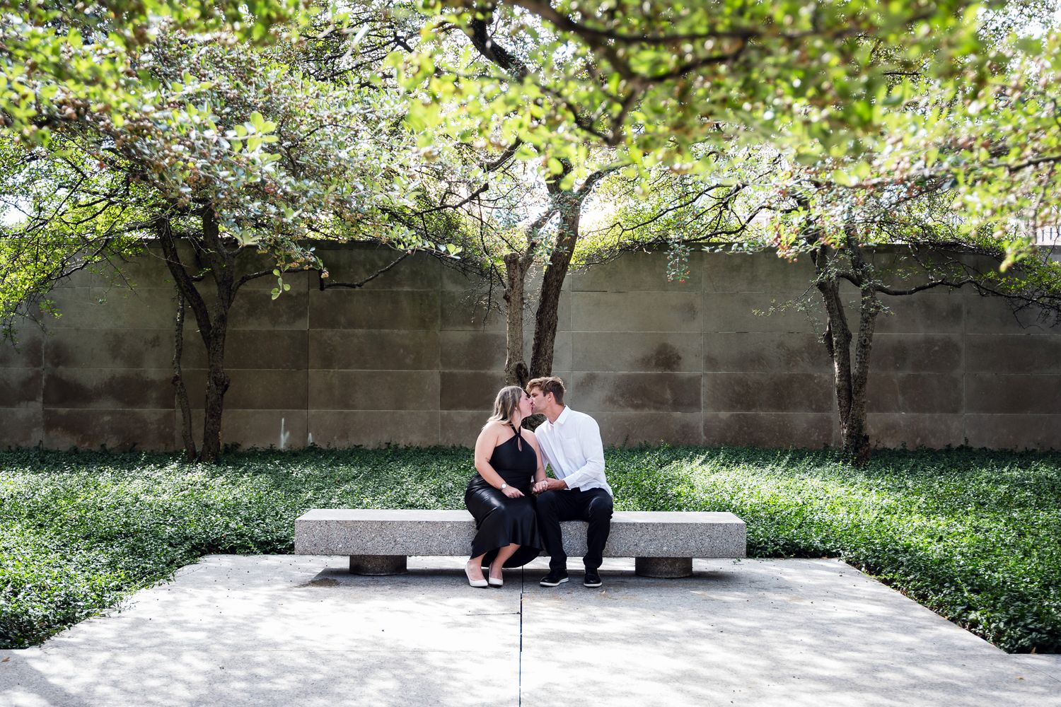 Couple sharing a romantic kiss on a stone bench in a serene garden courtyard with lush green trees.