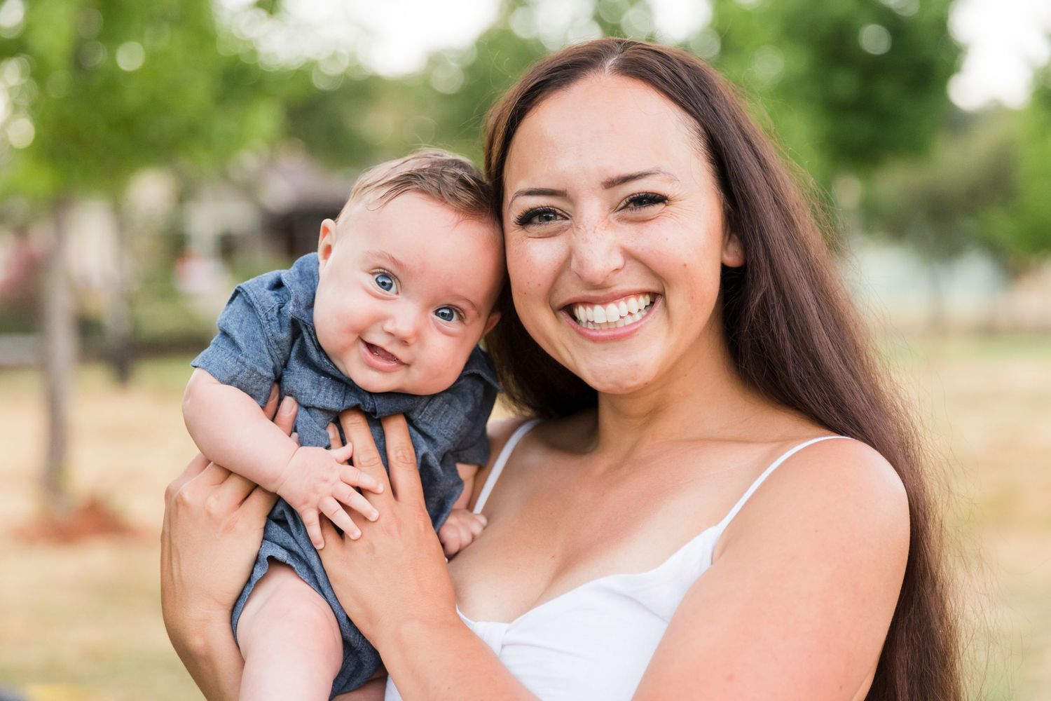 Smiling young woman holding a happy baby in a denim outfit outdoors in a green park setting.
