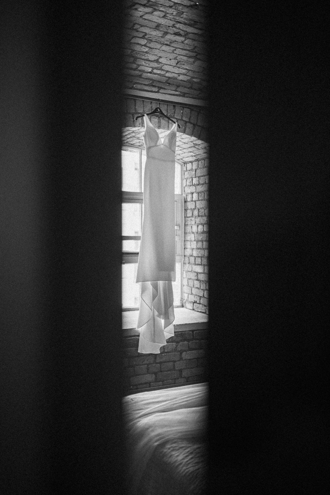 Black and white photo of a wedding dress hanging on a hanger in a brick-walled window.