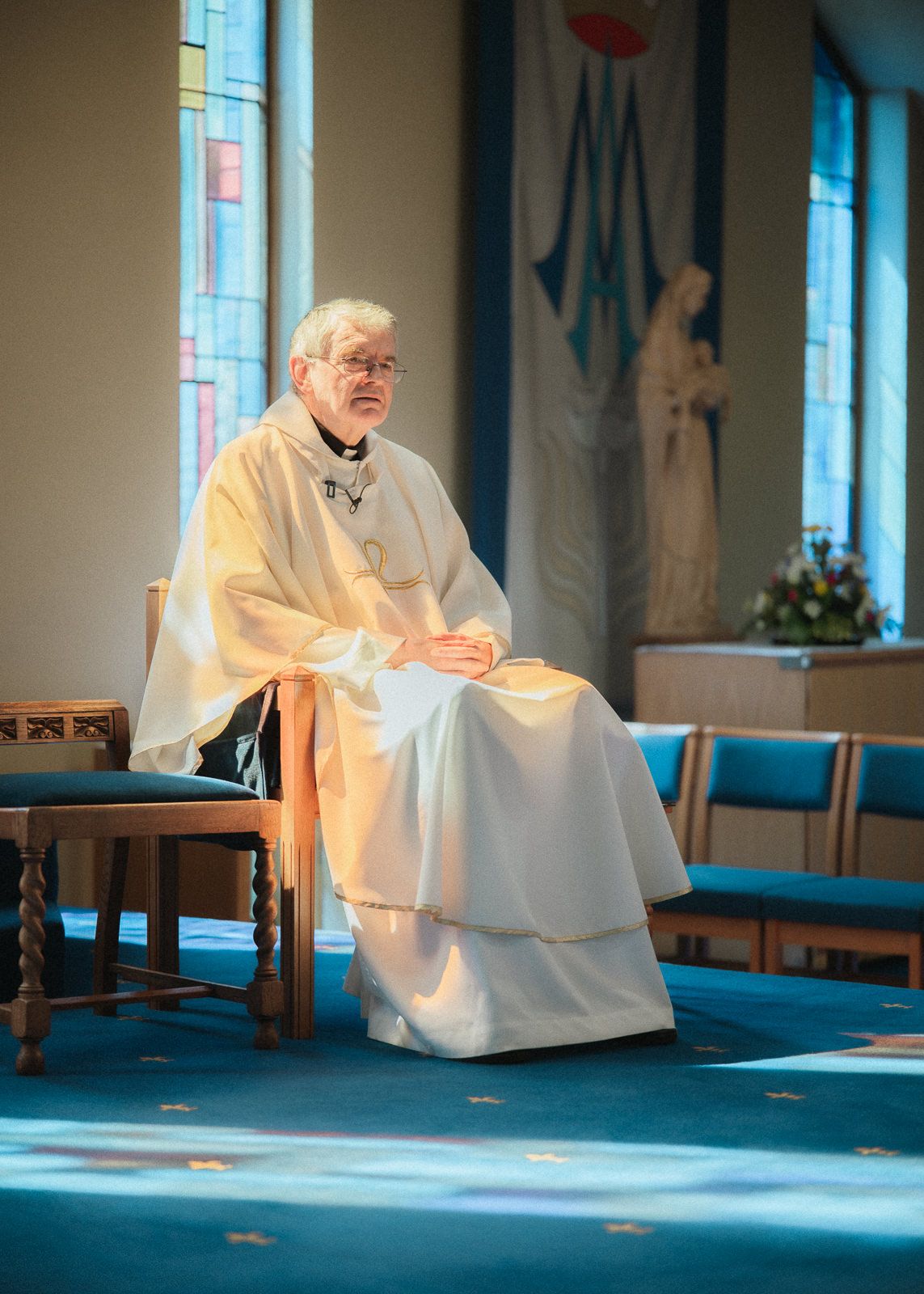A Catholic priest in white vestments sits quietly in a church with blue pews and stained glass windows.