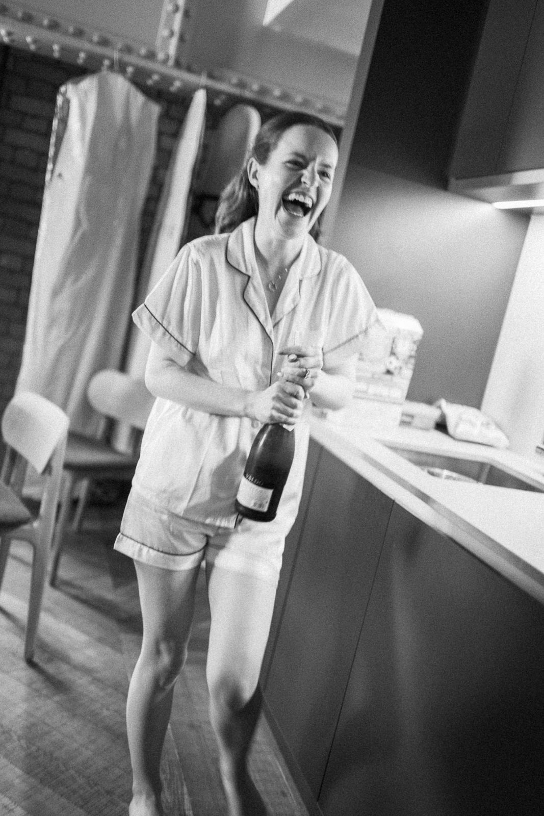 Black and white photo of a laughing woman in pajamas holding a champagne bottle in a modern kitchen.