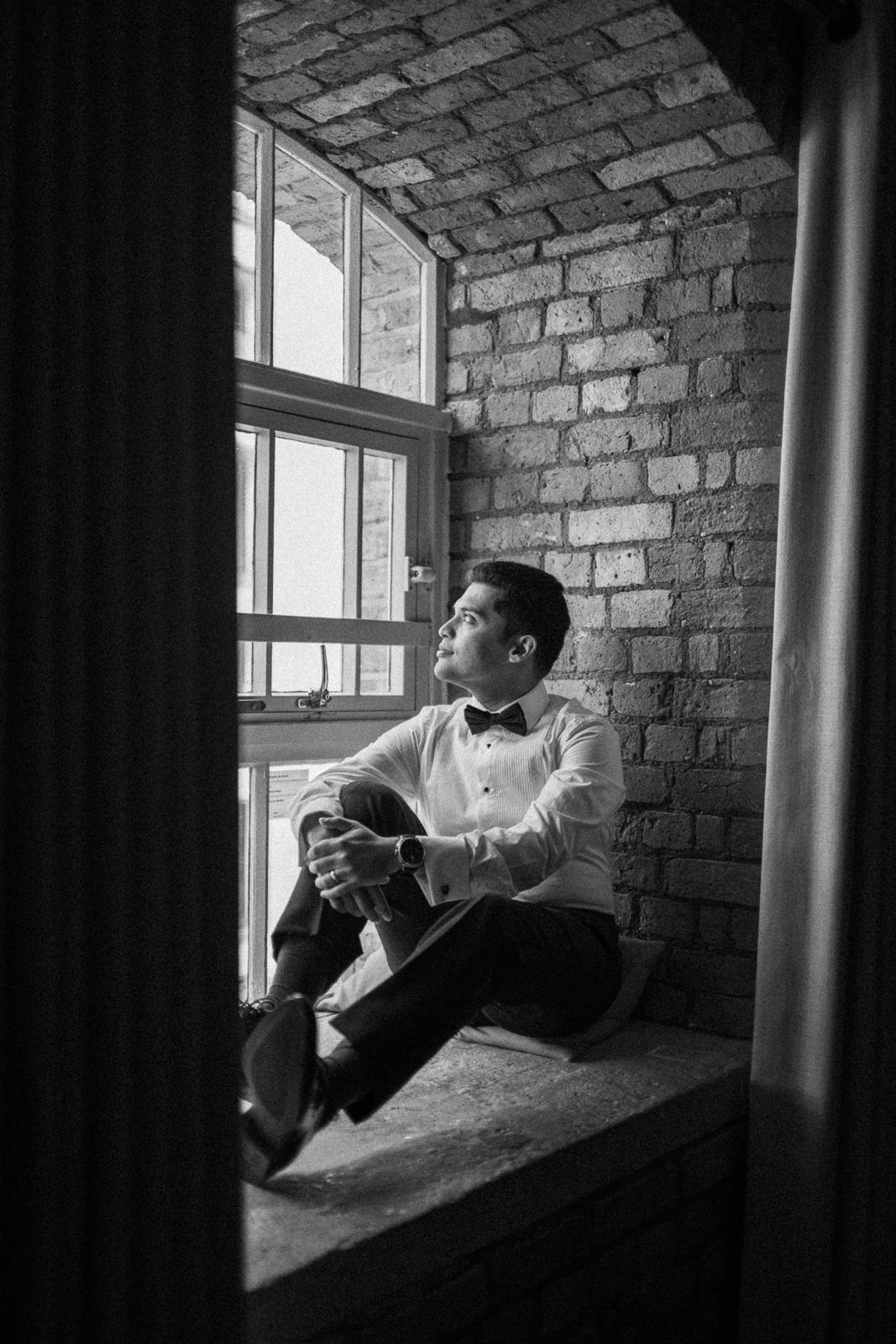 Black and white photo of a man in a tuxedo sitting on a window sill gazing outside against a brick wall.