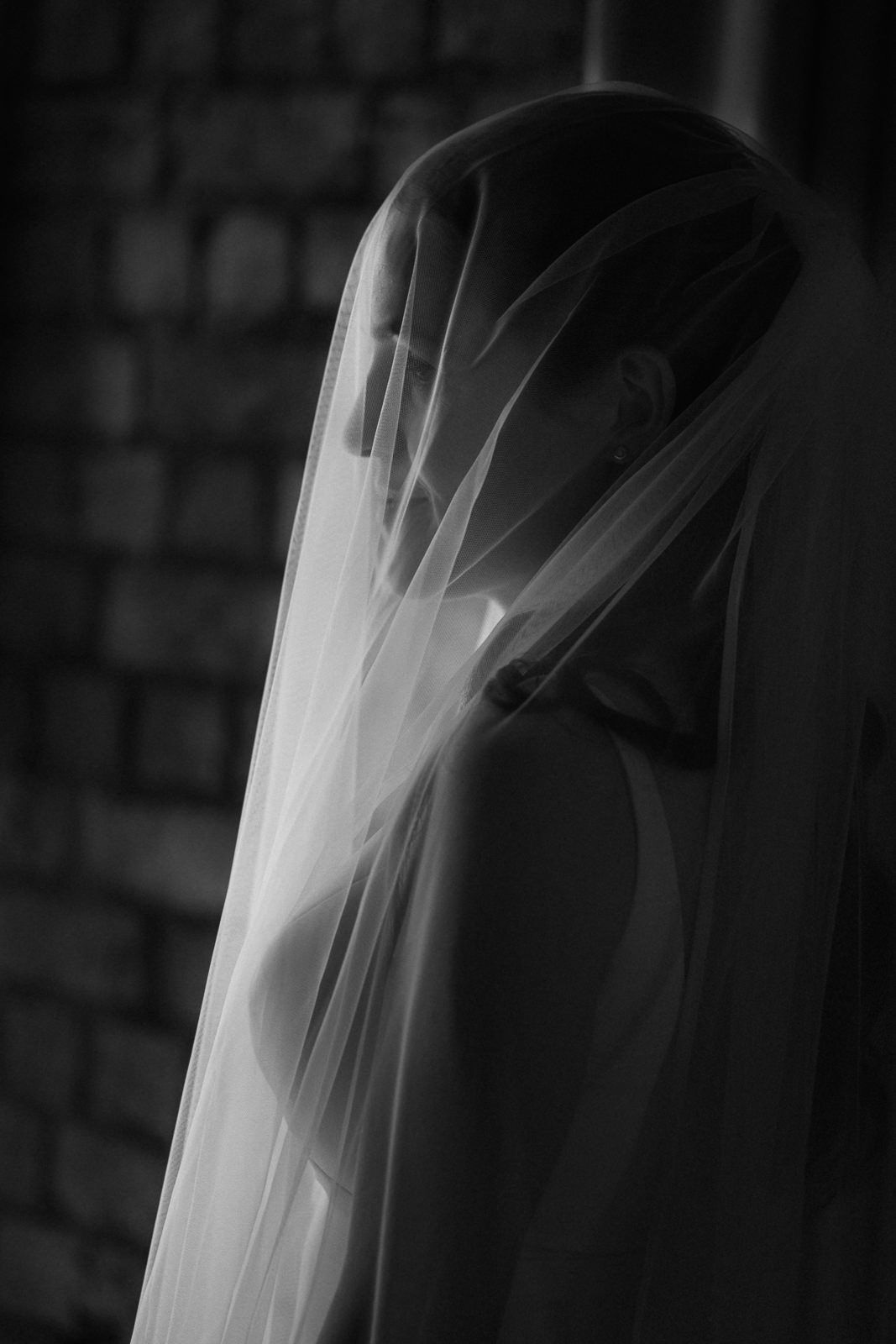 Black and white portrait of a bride covered by a flowing wedding veil against a brick wall.