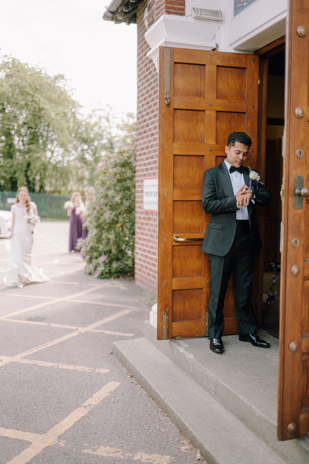 Groom in dark suit adjusting boutonniere at church door, with bridesmaids in purple dresses visible in background.