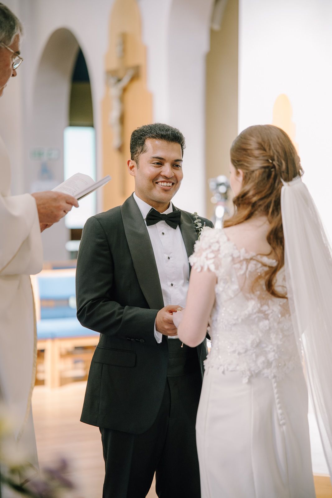 Groom in black tuxedo smiling at bride in lace wedding dress during church ceremony with officiant.