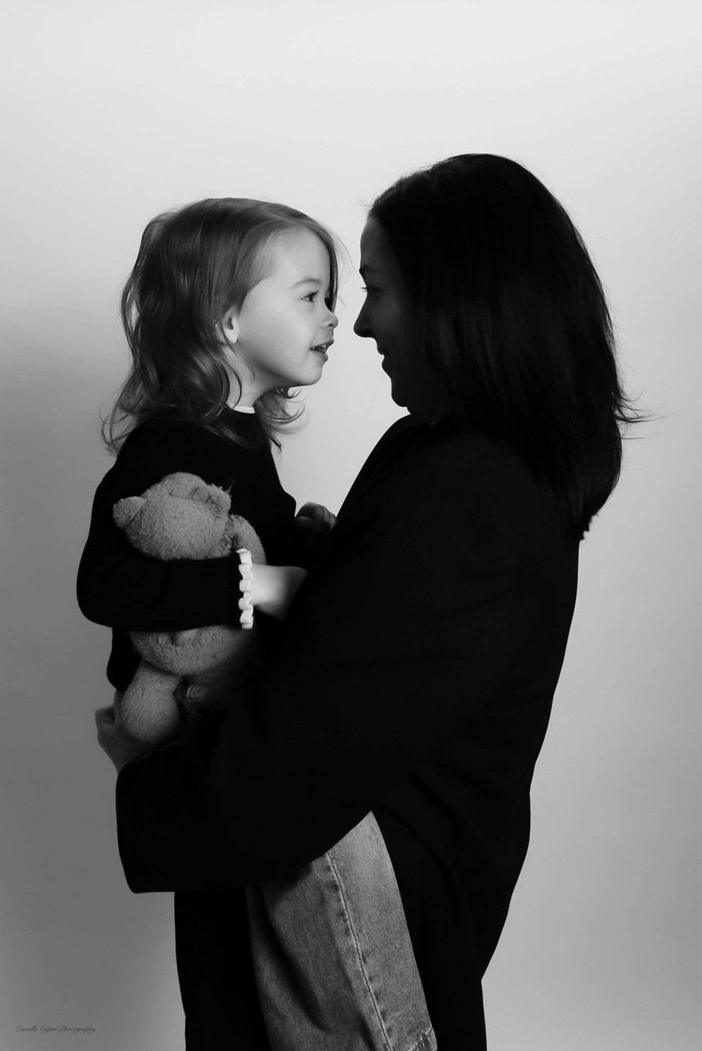 Black and white portrait of a mother holding her young daughter who clutches a teddy bear, sharing a tender moment, family portrait session