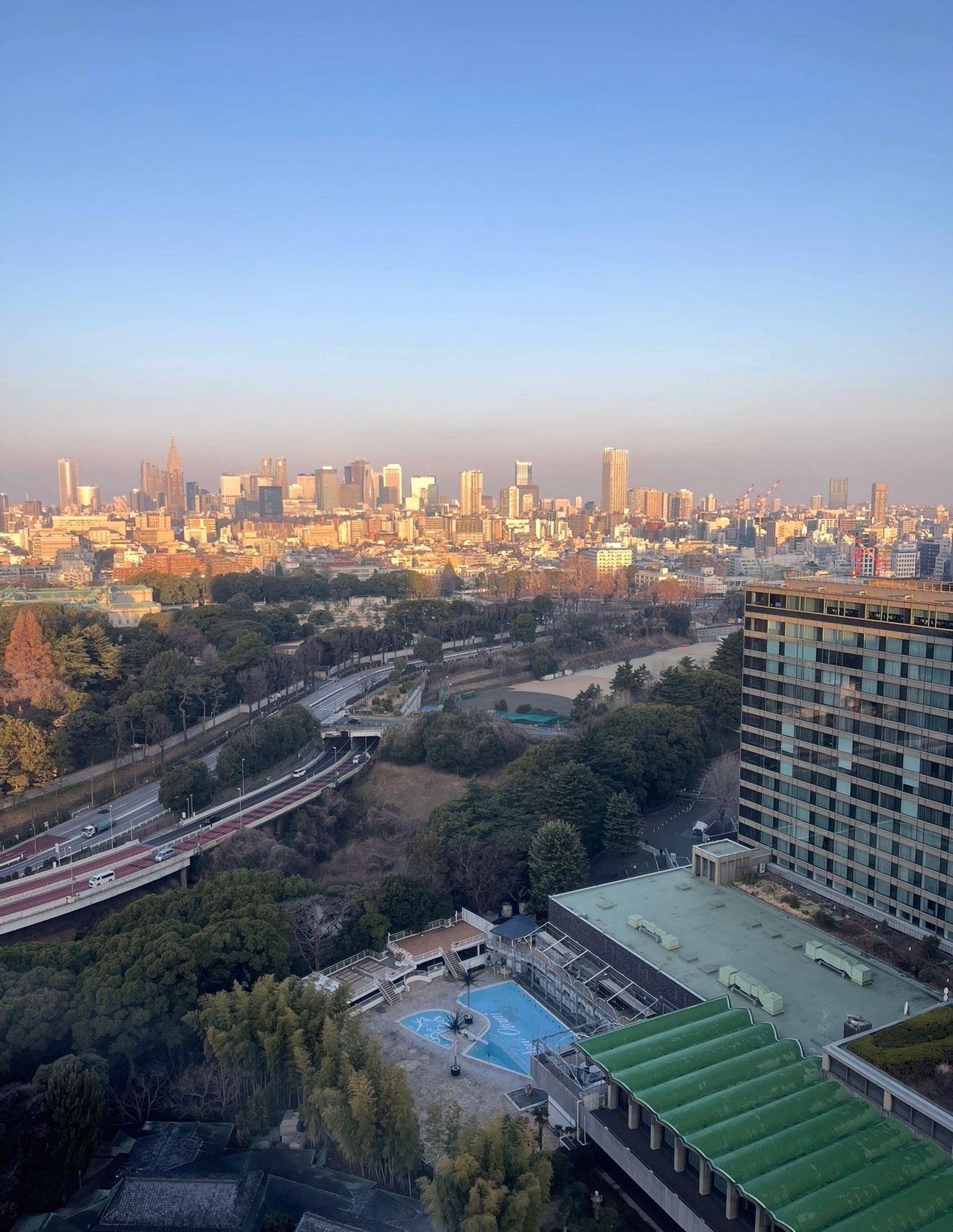 Aerial view of Tokyo skyline at golden hour with lush park, highway, and luxury hotel pool in foreground.
