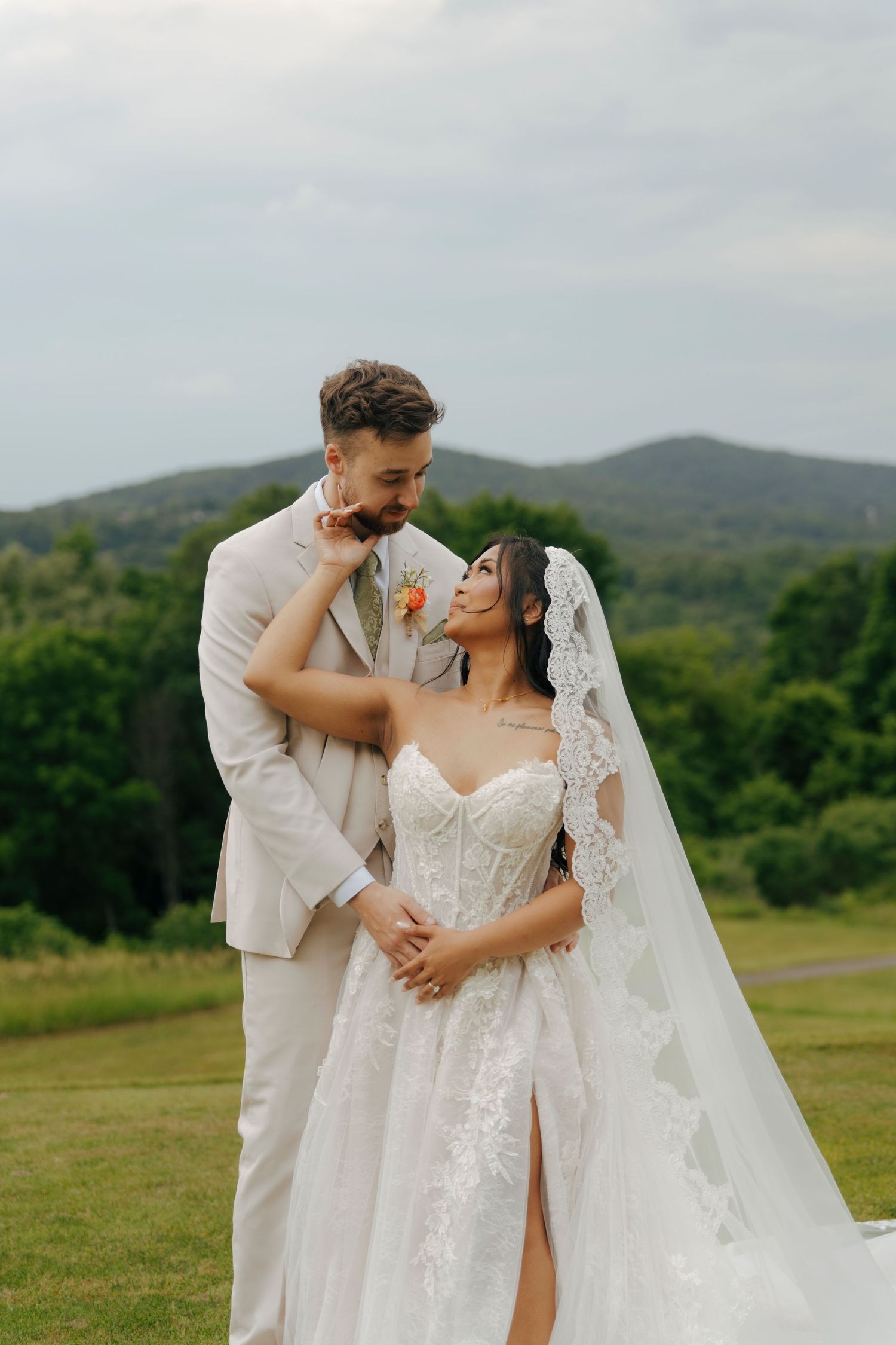 Bride in lace gown and veil with groom in beige suit posing outdoors with green hills backdrop.