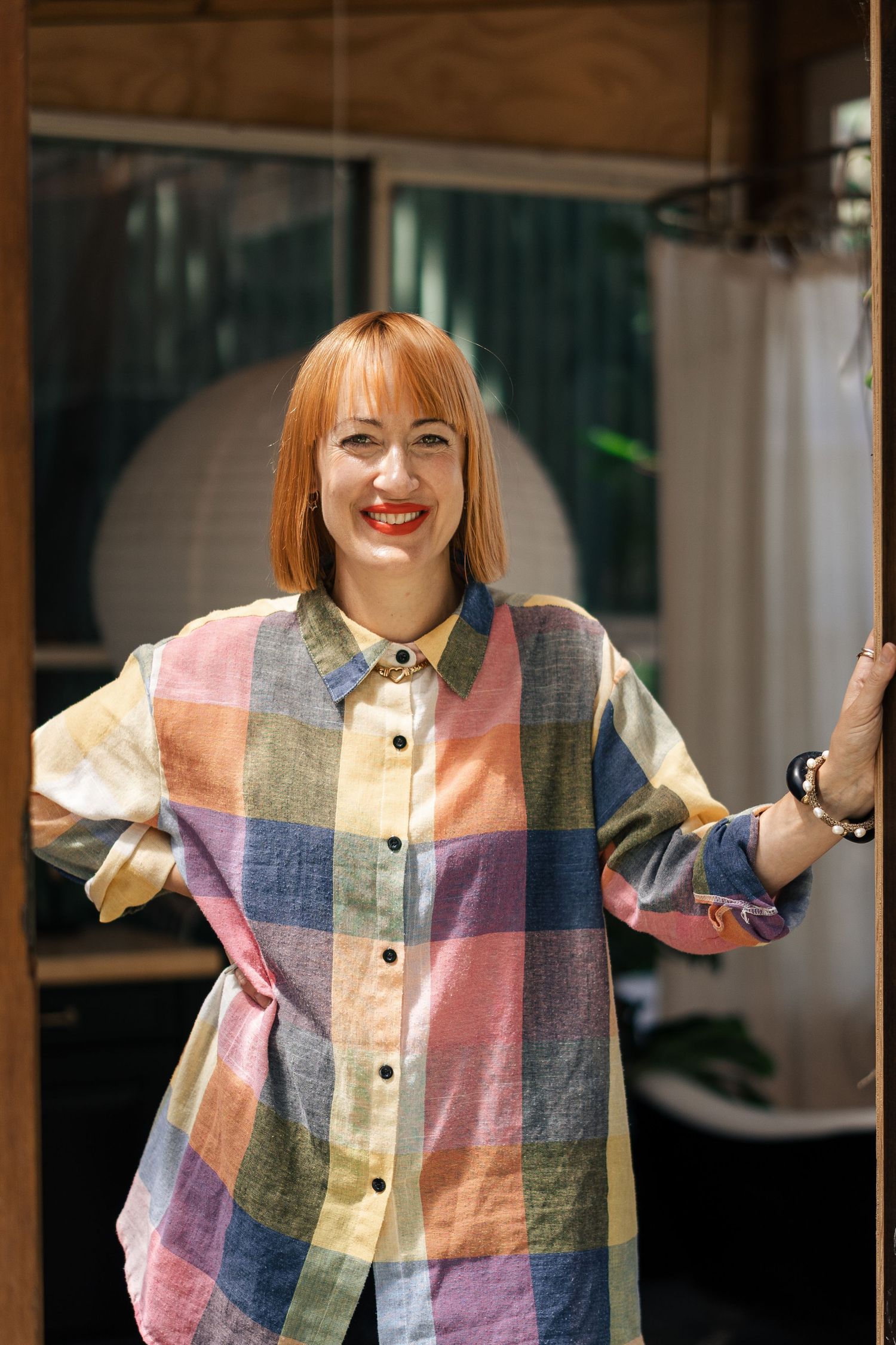 A cheerful redhead woman in a colorful plaid oversized shirt poses in a sunlit doorway.

Mimi Moon Studio