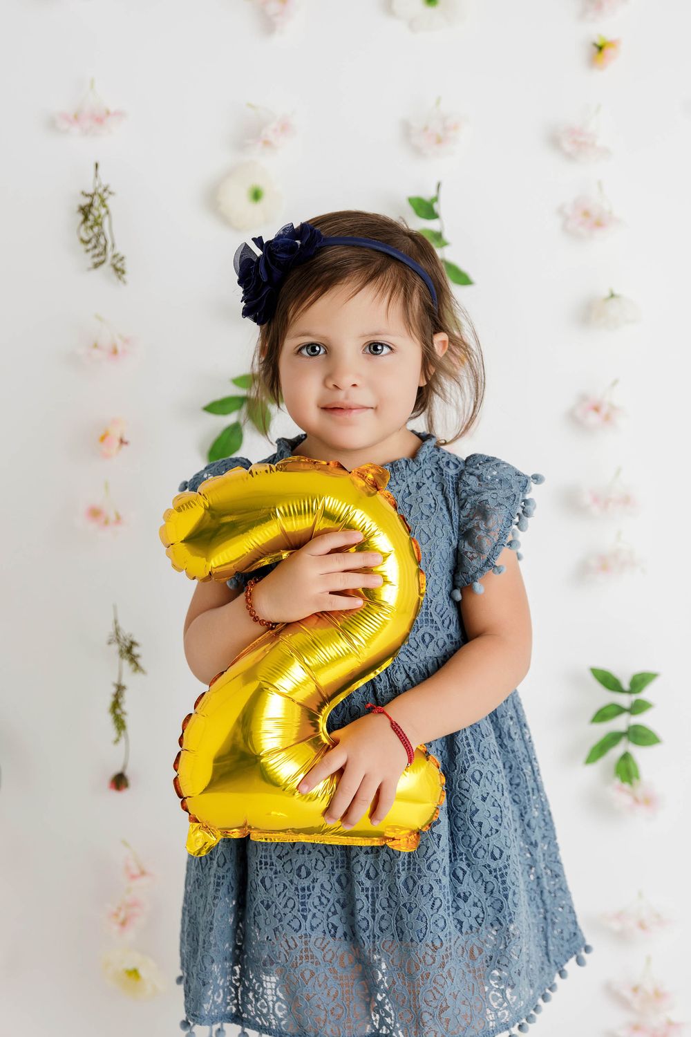 A toddler girl in a blue lace dress holds a gold number 2 balloon, celebrating her 2nd birthday.