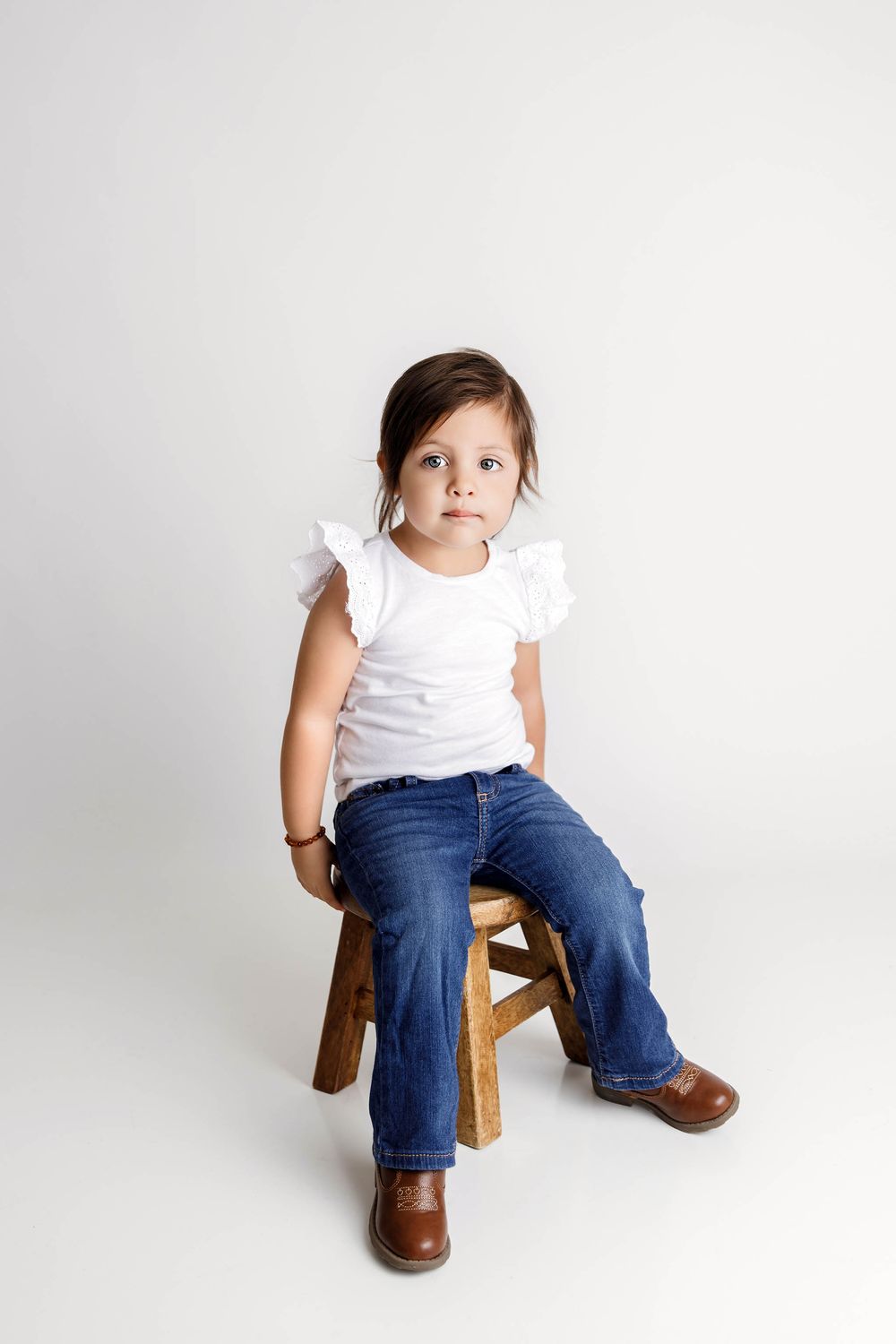 A cute toddler girl in white top and jeans sits on a wooden stool against a white background.