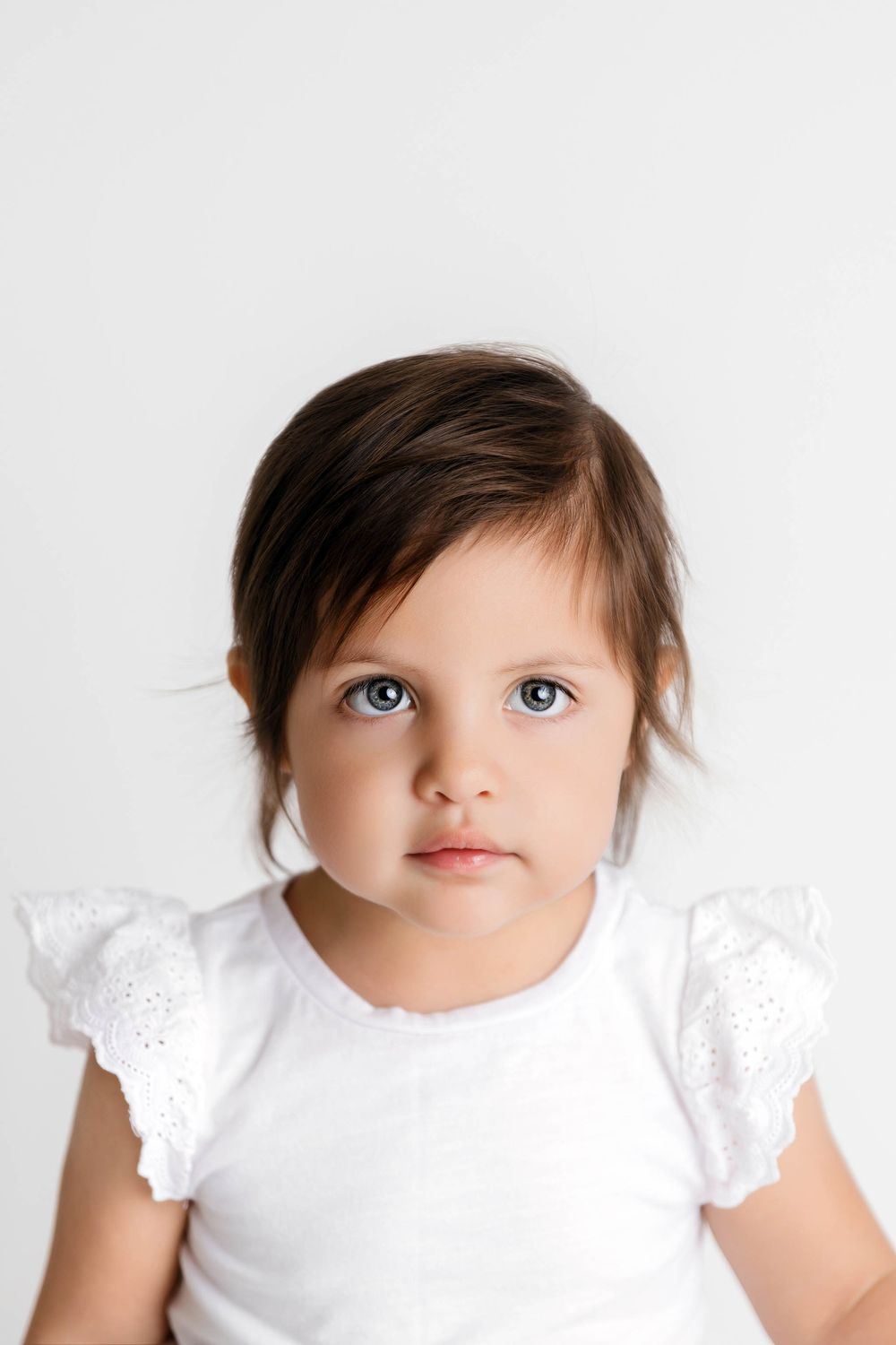 Adorable toddler girl with dark hair and blue eyes wearing a white ruffle top against a white background.