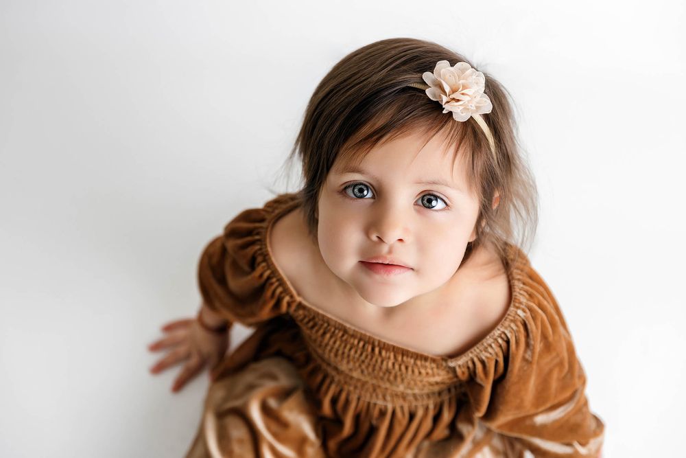 Adorable toddler girl in brown fringe dress with floral headband looking up, studio portrait on white background.