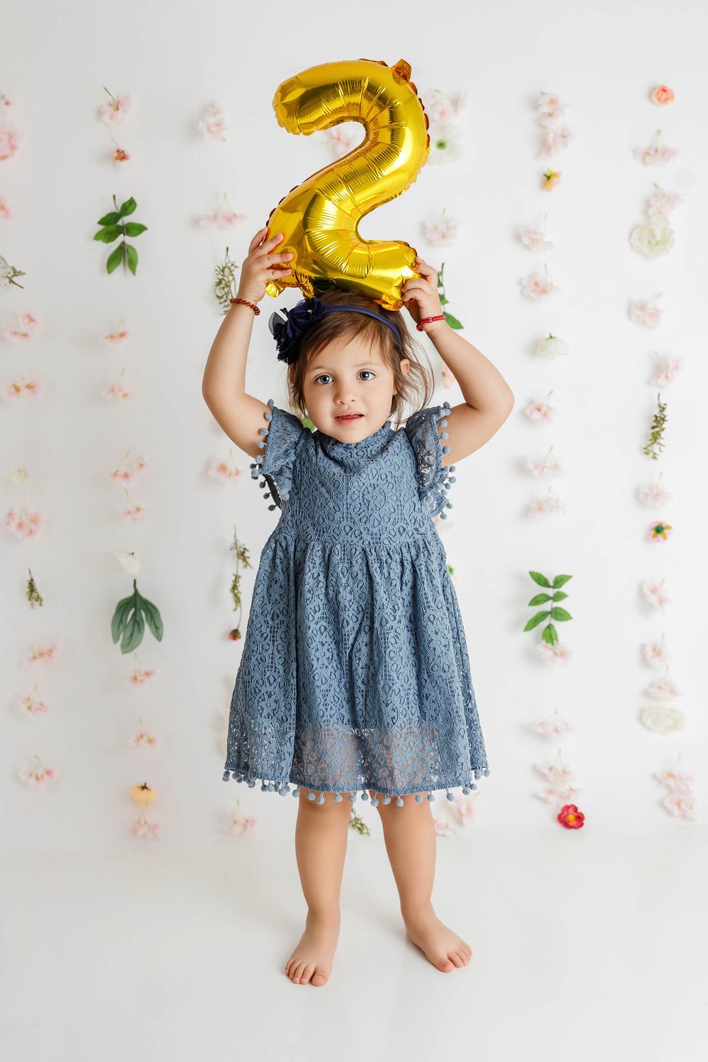 A toddler girl in a blue lace dress holds a gold number 2 balloon above her head against a floral white backdrop.