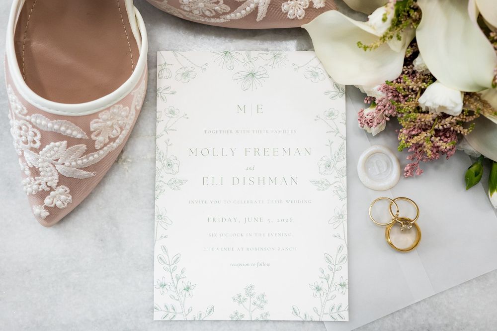 Wedding invitation for Molly Freeman and Eli Dishman displayed with lace bridal heels, gold rings, and white floral bouquet.