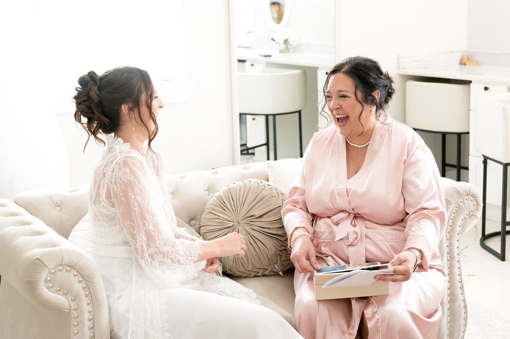 Bride in white lace gown and woman in pink satin robe share a joyful moment on an elegant cream sofa.