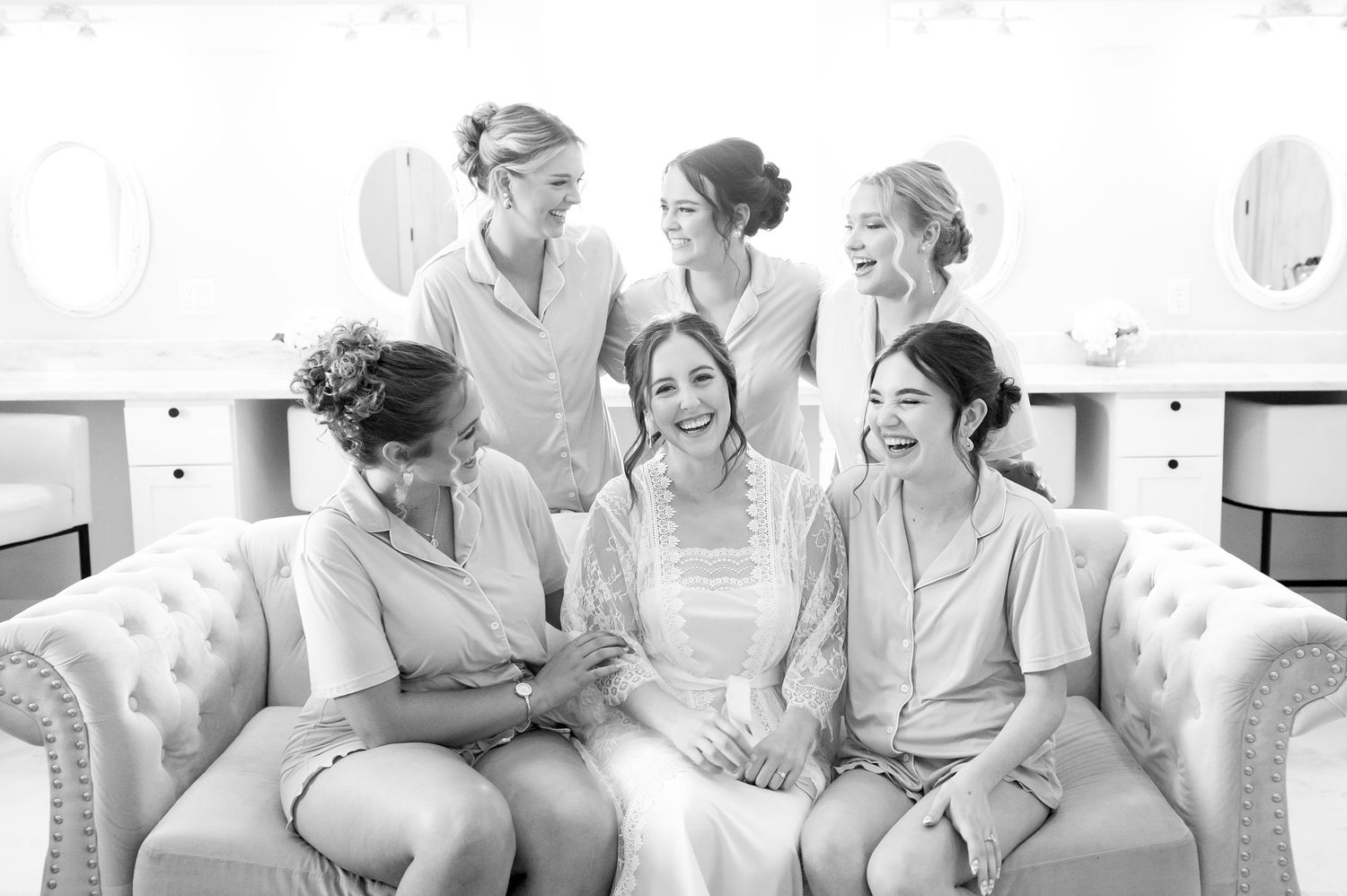 Bride surrounded by smiling bridesmaids and stylists on tufted sofa during wedding day preparations.