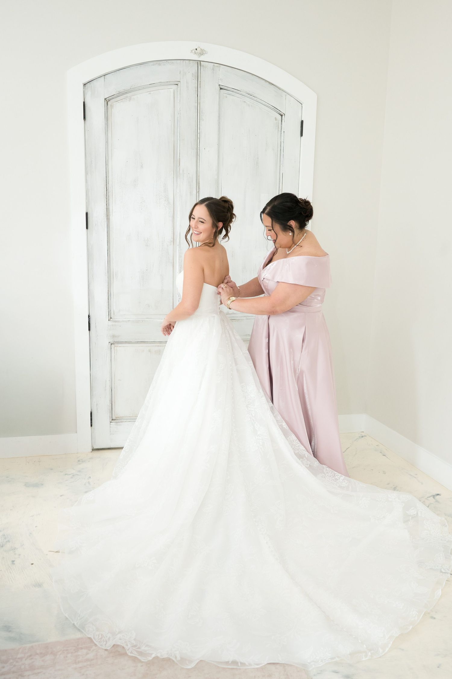 Bridesmaid in blush pink dress helping bride button her elegant white ball gown wedding dress before ceremony.
