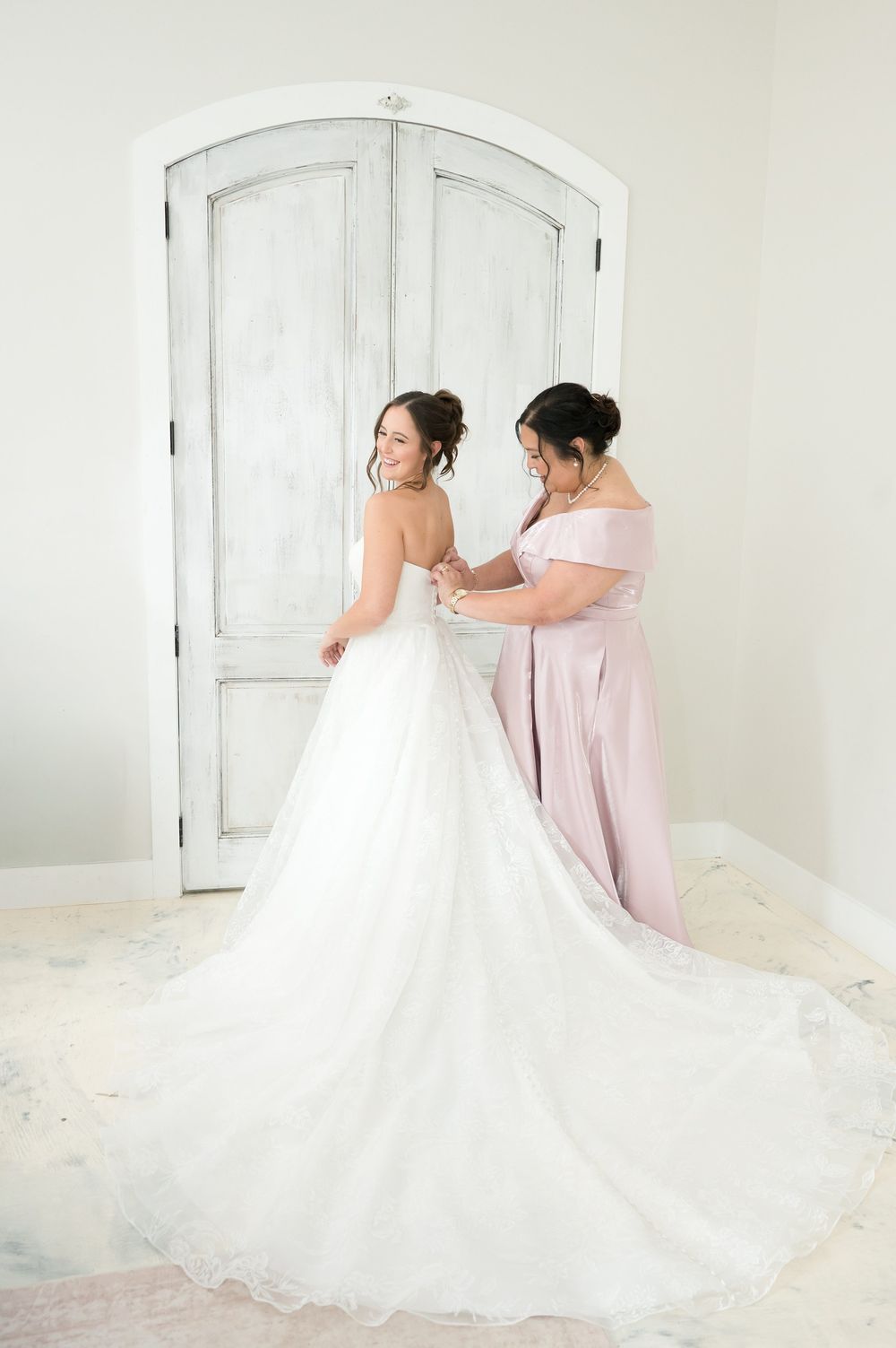 Bridesmaid in blush pink dress helping bride button her elegant white ball gown wedding dress before ceremony.