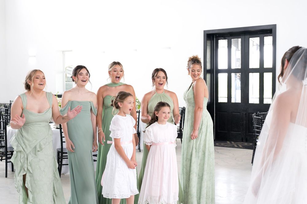 Bridesmaids in sage green dresses react with joy seeing the bride, alongside flower girls in white dresses.