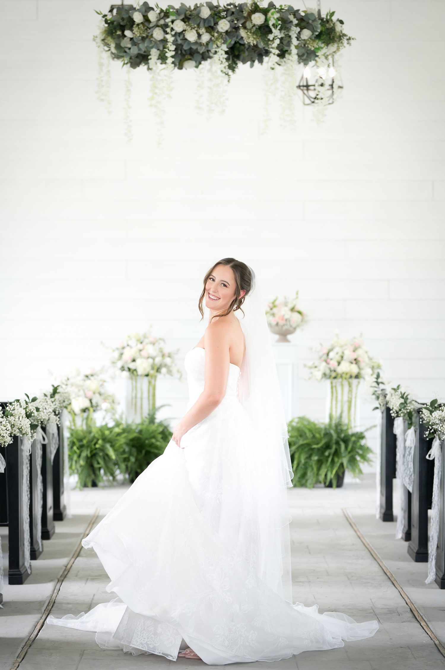 Bride in white strapless gown twirls in elegant chapel aisle with lush floral arrangements and greenery overhead.