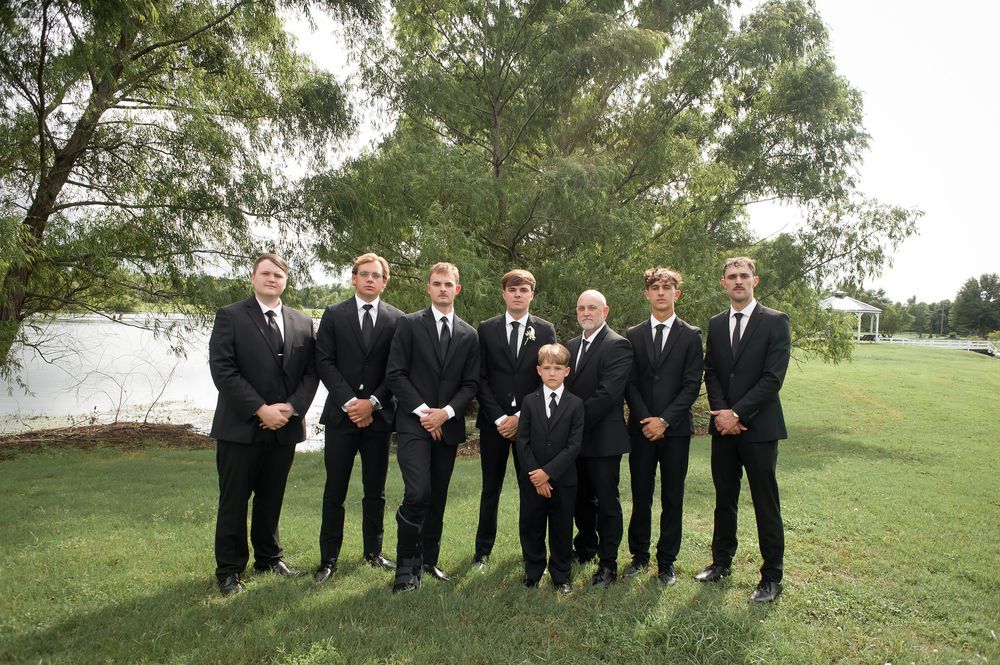 Seven groomsmen in matching black suits pose outdoors on green grass with trees in the background.