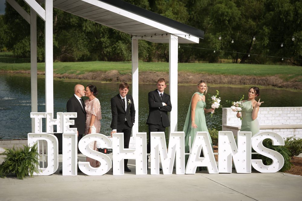 Wedding party posing beside large white THE DISHMANS letter sign near a scenic waterfront pavilion.