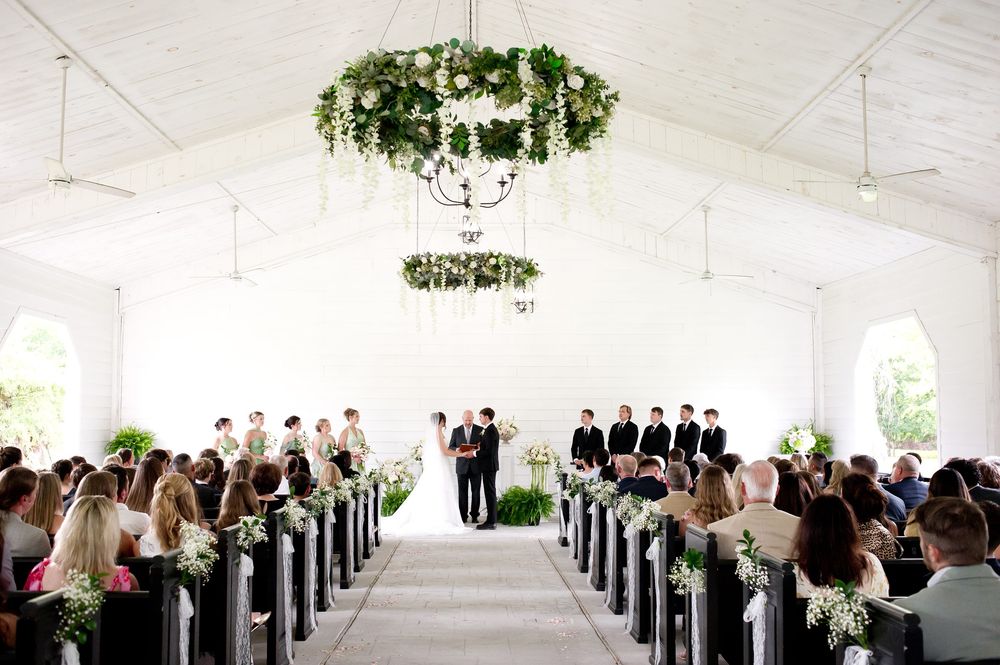 Elegant wedding ceremony in a white tent venue with floral chandeliers, pew decorations, and guests seated along a classic aisle.
