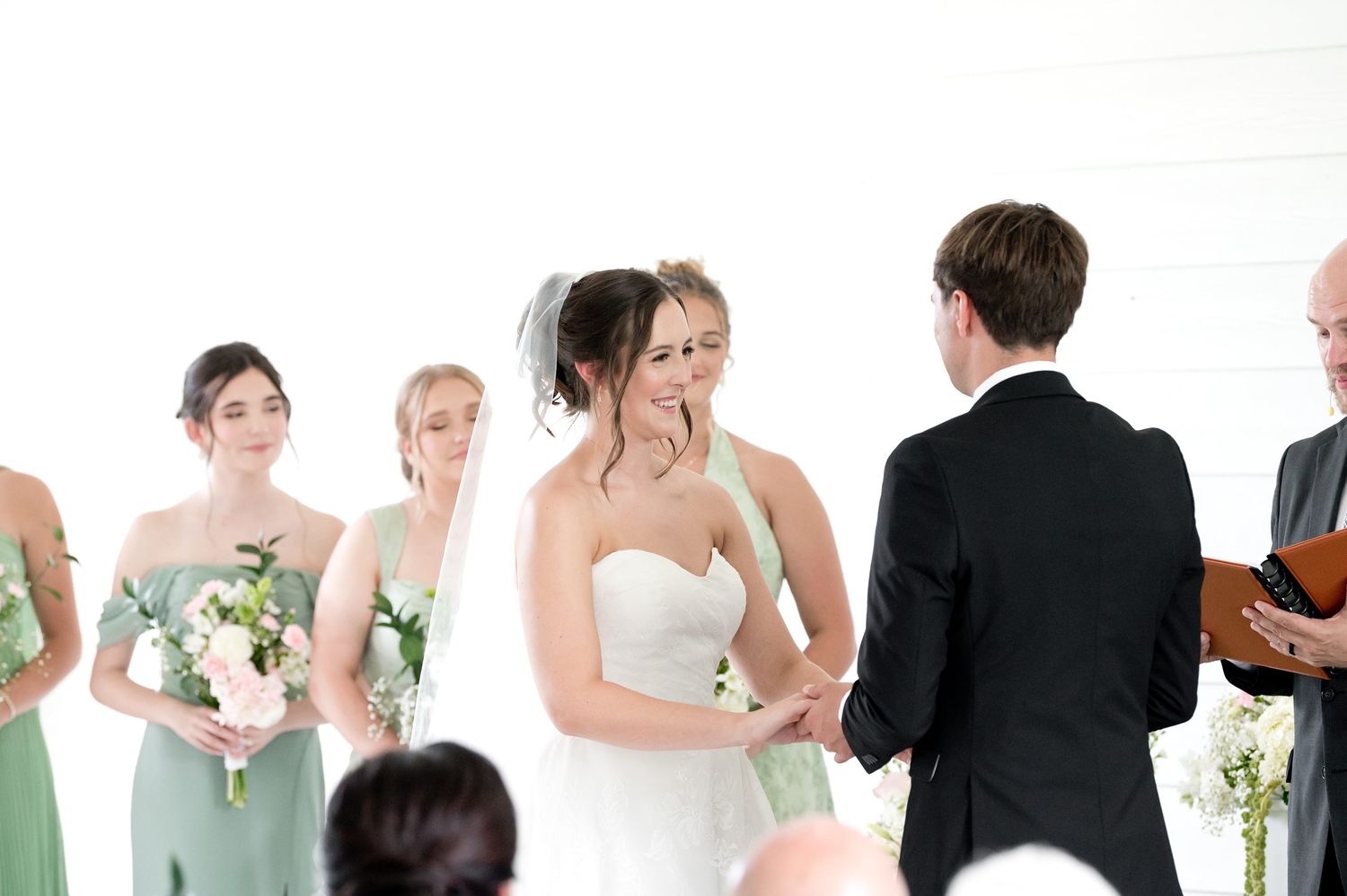 Bride smiling at groom during wedding ceremony with bridesmaids in sage green dresses holding white floral bouquets.