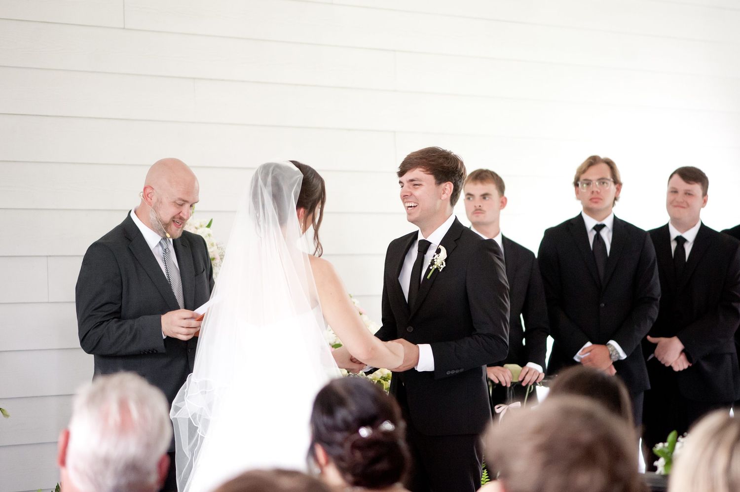 Wedding ceremony with groom holding bride's hands before officiant, groomsmen standing in background at elegant white venue.