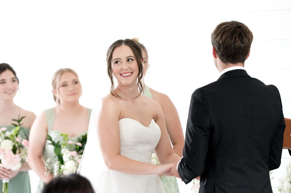 Bride smiling at groom during wedding ceremony with bridesmaids in sage green dresses holding pink bouquets.