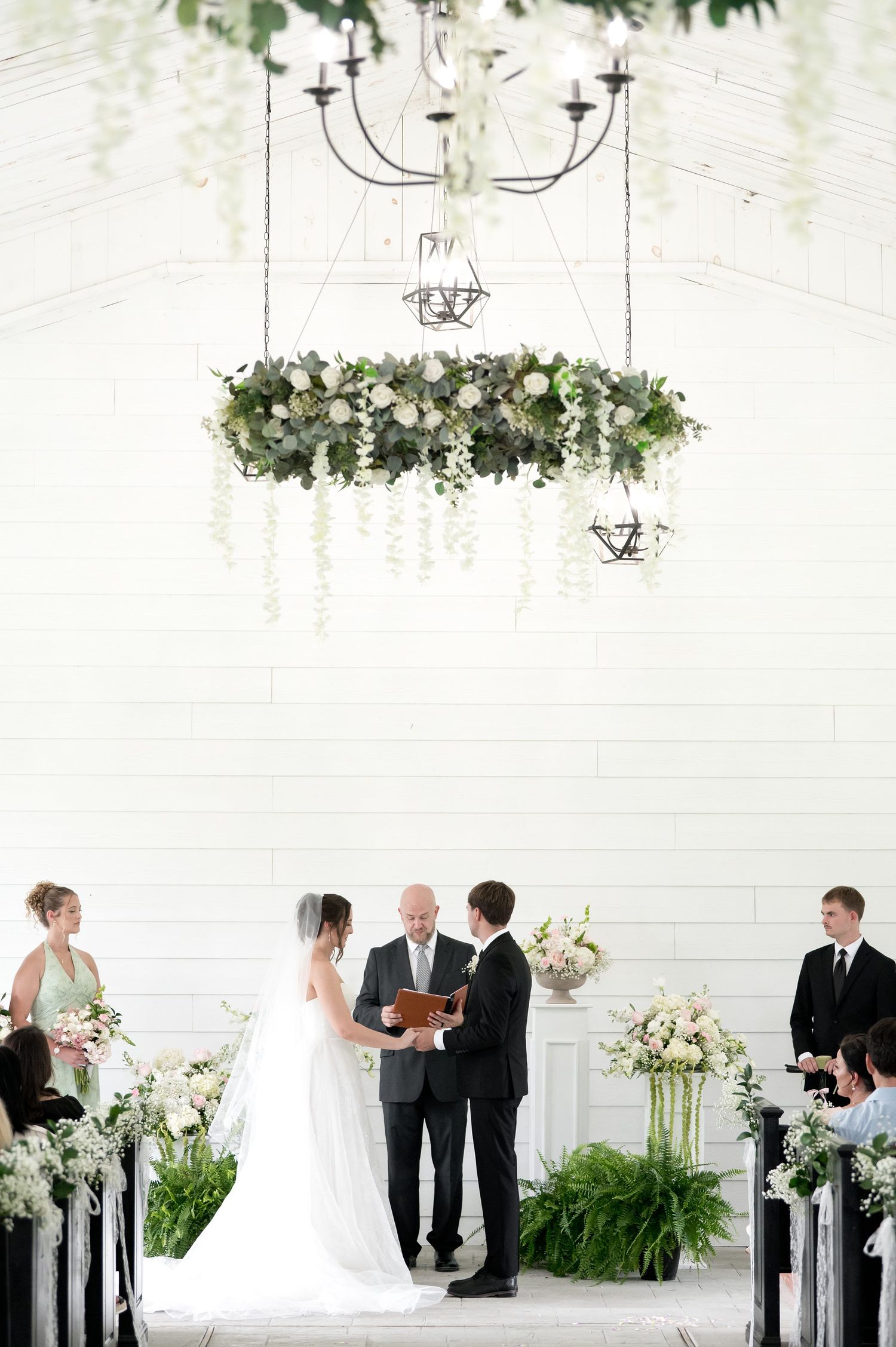 Elegant wedding ceremony in white chapel with floral chandelier, greenery, and white flowers as couple exchanges vows at altar.