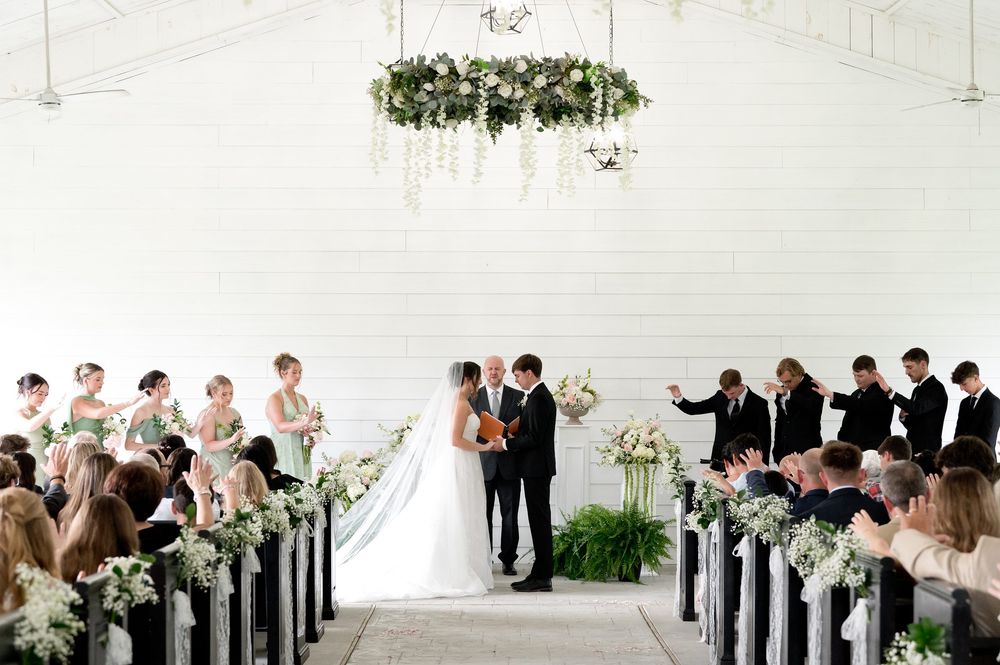 Elegant wedding ceremony in a white chapel with floral chandelier, bridal party, and guests seated in pews.