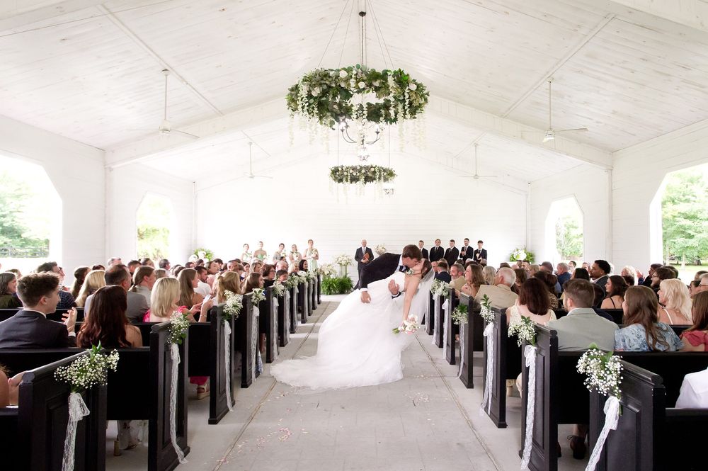 Elegant white chapel wedding ceremony with wooden pews, floral chandelier, and bride walking down the aisle.