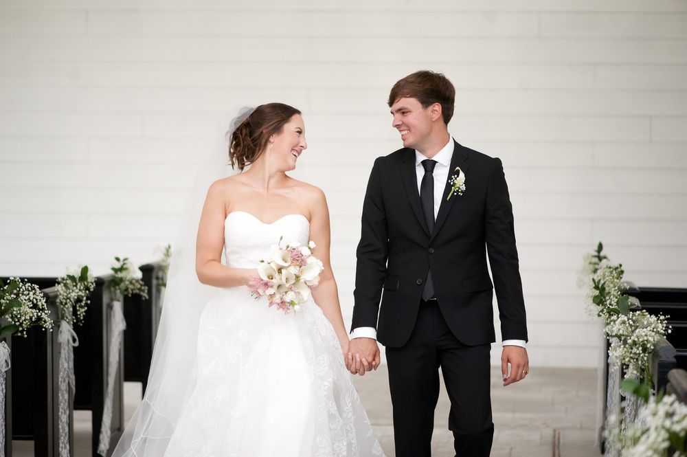 Bride in white gown holding bouquet and groom in black suit hold hands smiling at wedding ceremony.