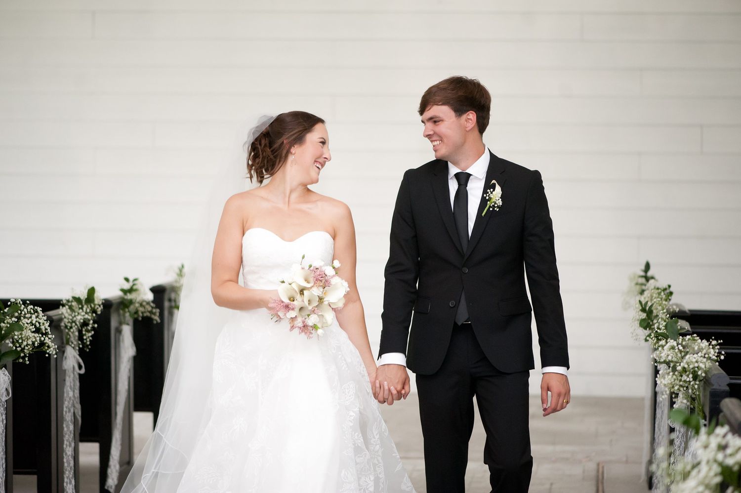 Bride in white gown holding bouquet and groom in black suit hold hands smiling at wedding ceremony.