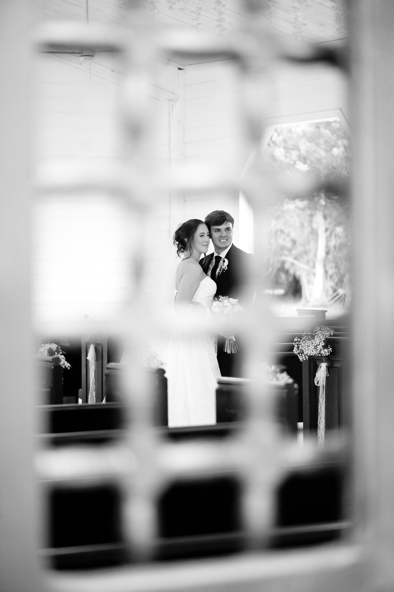 Black and white wedding photo of bride and groom embracing, captured through a decorative window frame.