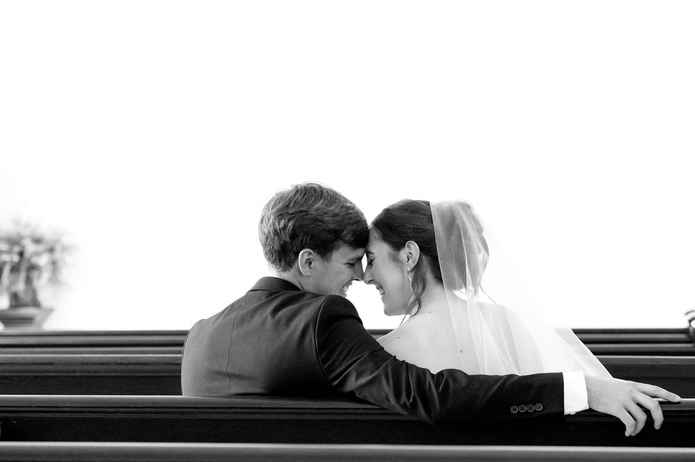 Romantic black and white photo of bride and groom sitting closely together on a church pew.