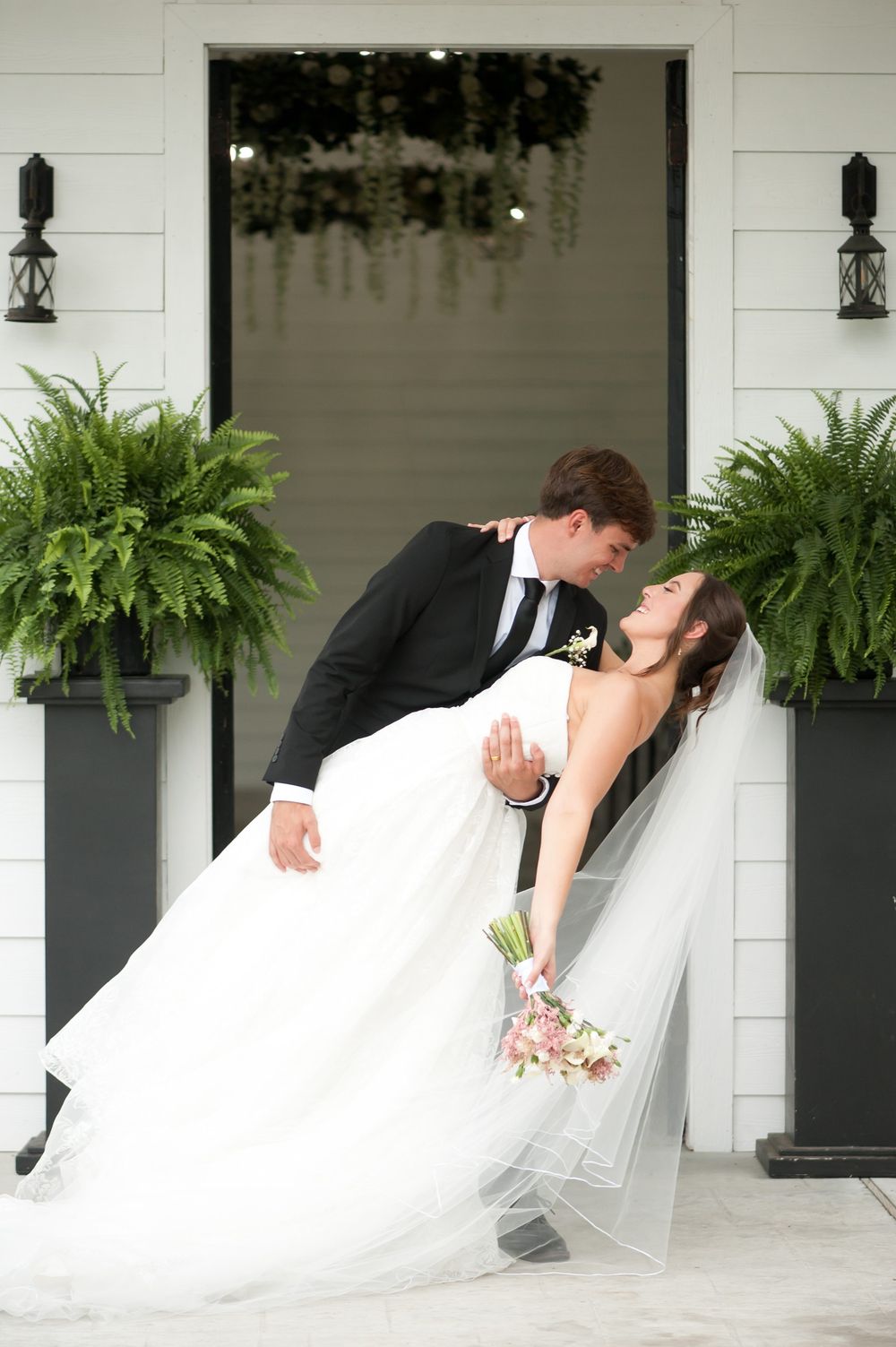 Groom dips bride in white gown and veil outside elegant white venue with ferns and floral chandelier.