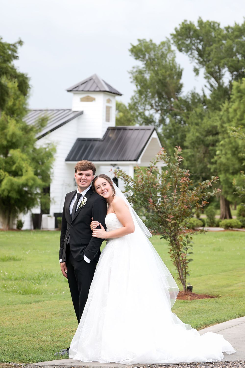 Bride and groom posing together outdoors on green lawn with white chapel in background on wedding day.