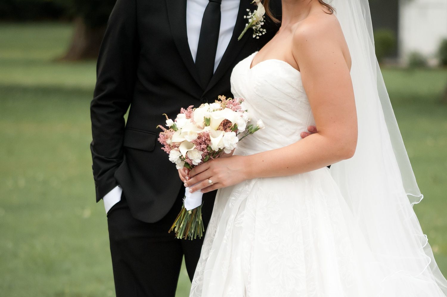 Bride and groom standing together outdoors, bride holding a white and pink floral bouquet in a strapless wedding gown.