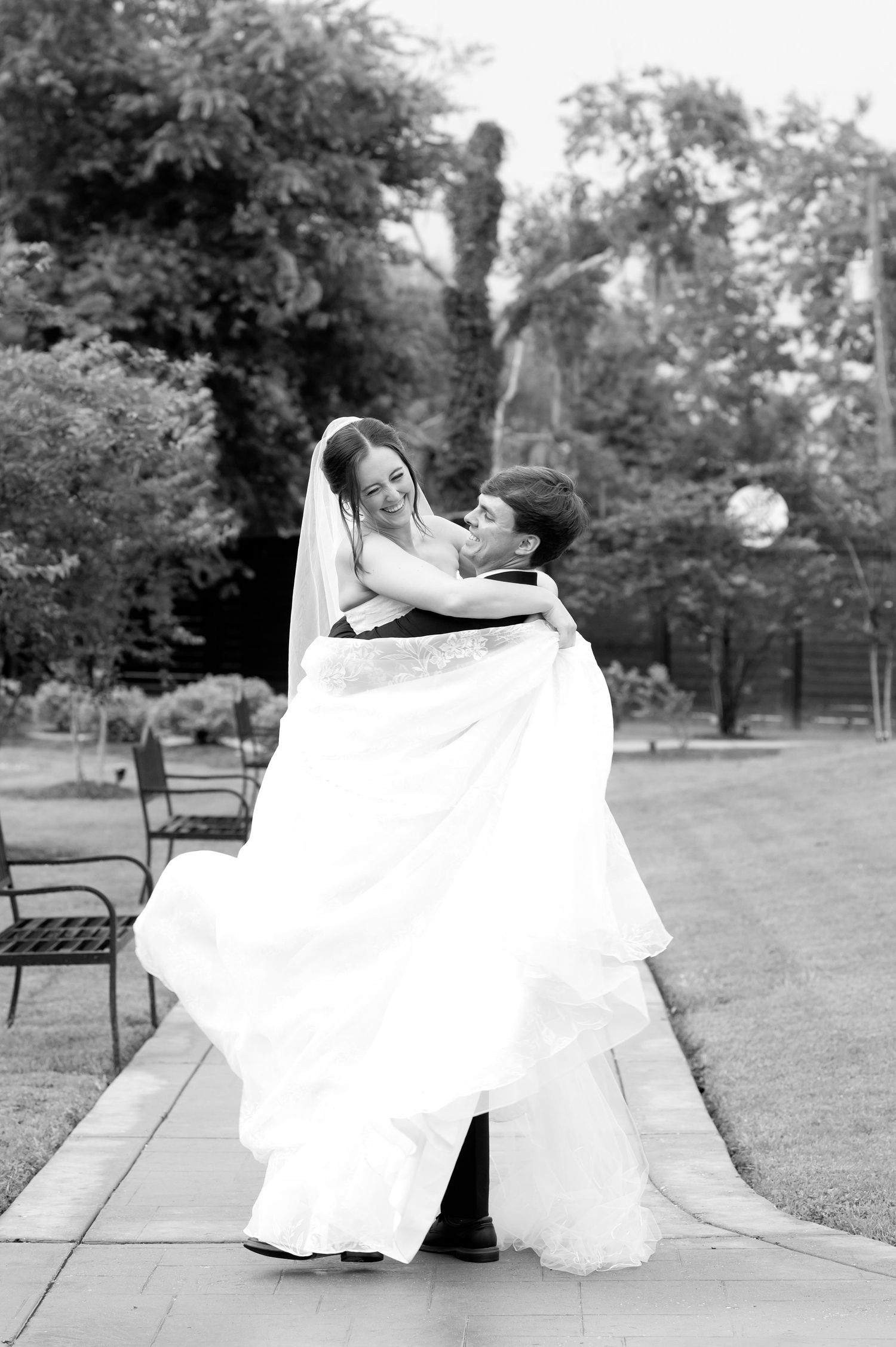 Groom lifting bride in white wedding gown and veil outdoors on a garden pathway in black and white photo.