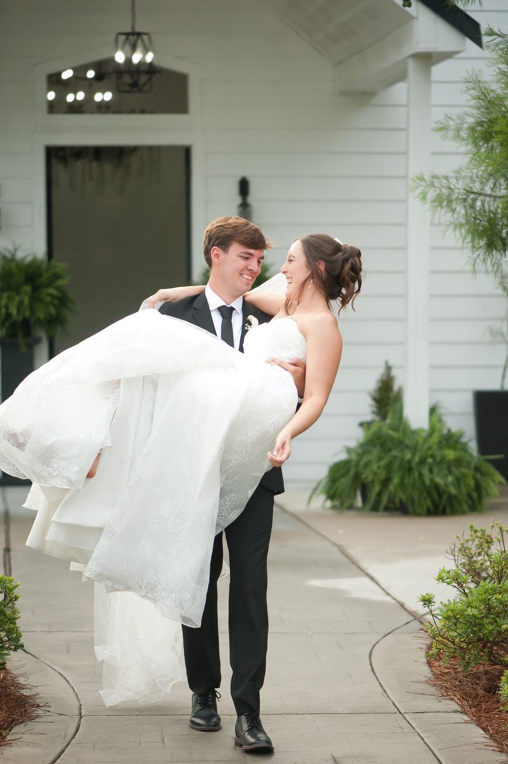 Groom carrying bride in white wedding gown outside white venue, sharing a joyful romantic moment.