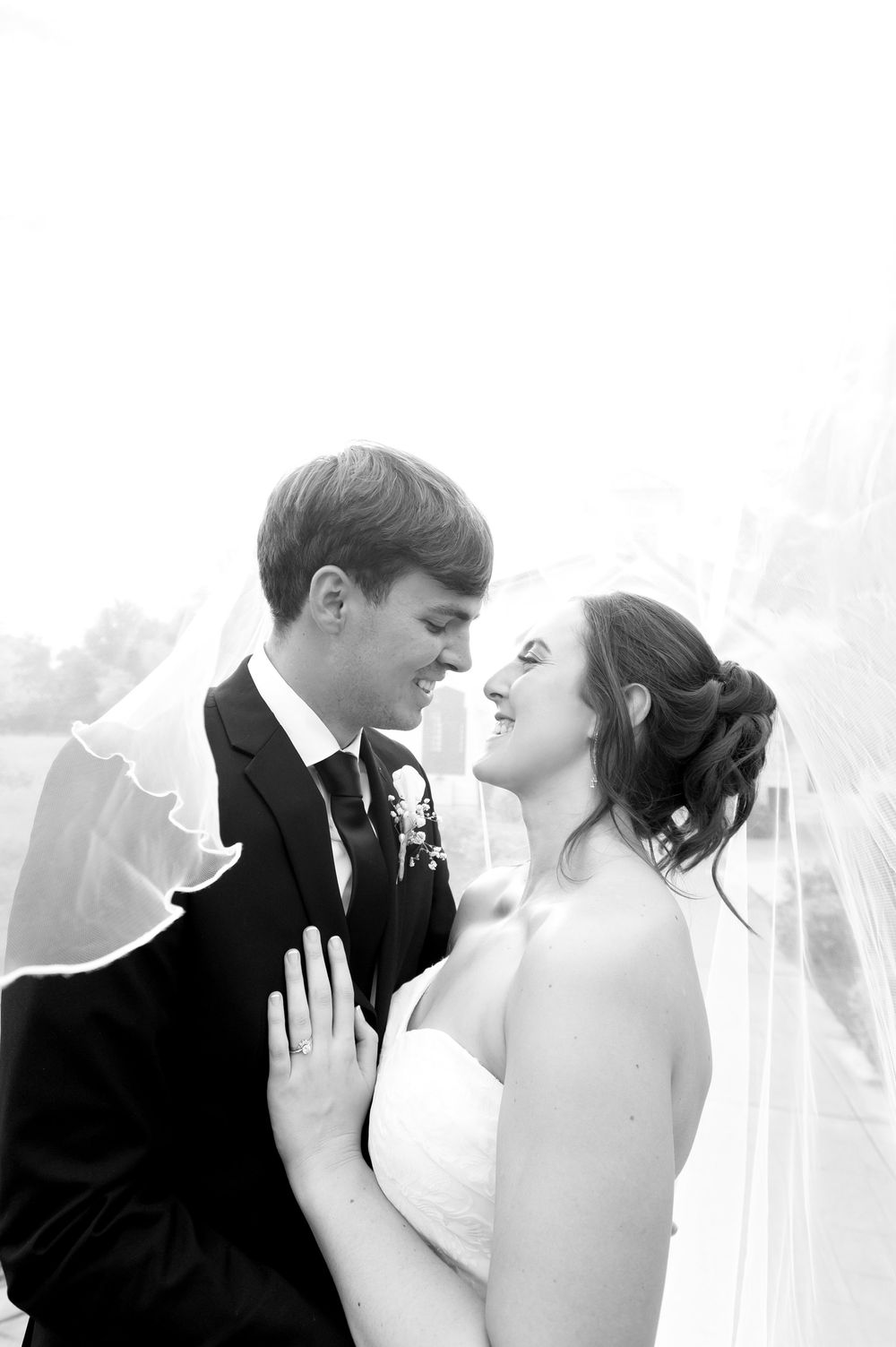 Black and white wedding photo of smiling bride and groom gazing at each other under flowing veil.