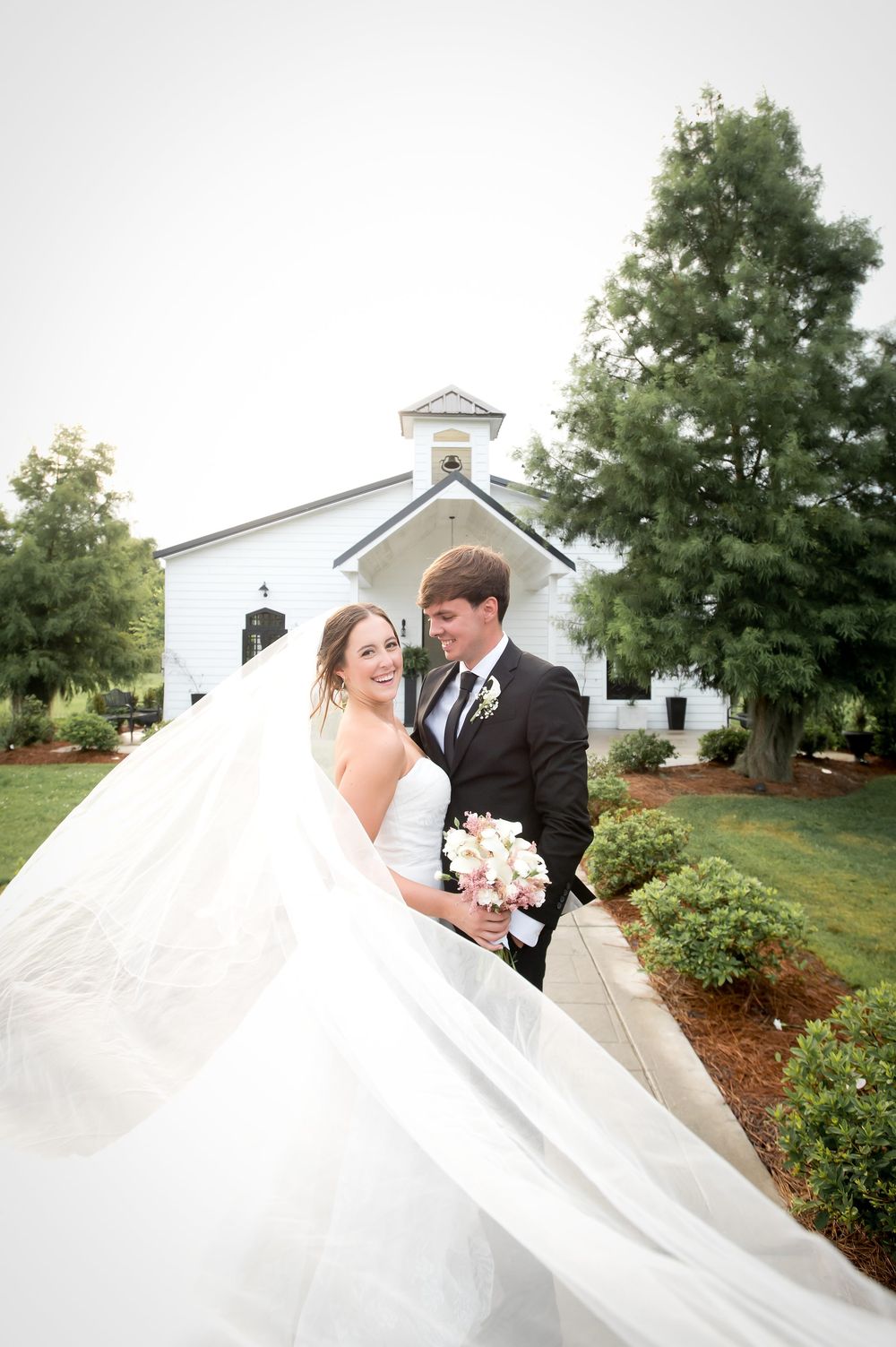 Bride and groom pose in front of a white chapel with veil flowing in the wind on their wedding day.