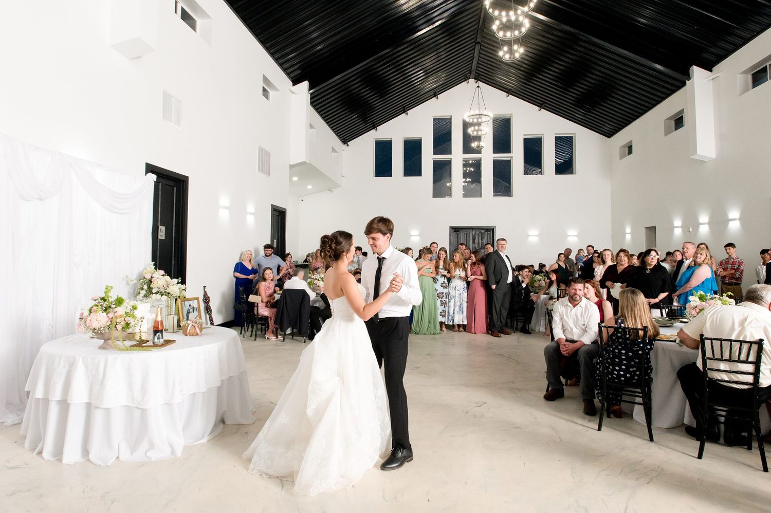 Bride and groom share first dance in a white vaulted chapel reception hall surrounded by seated wedding guests.